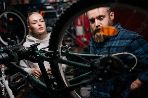 Cycling mechanic male with beard communicating with attractive blonde female client, talking problem of bicycle, detected during diagnostics in repair shop with dark interior, standing behind MTB.