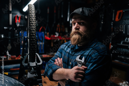 Side view of bearded cycling repairman in cap holding wrench in hand standing by bicycle in repair workshop with dark interior, serious looking at camera. Concept of professional bicycle maintenance.