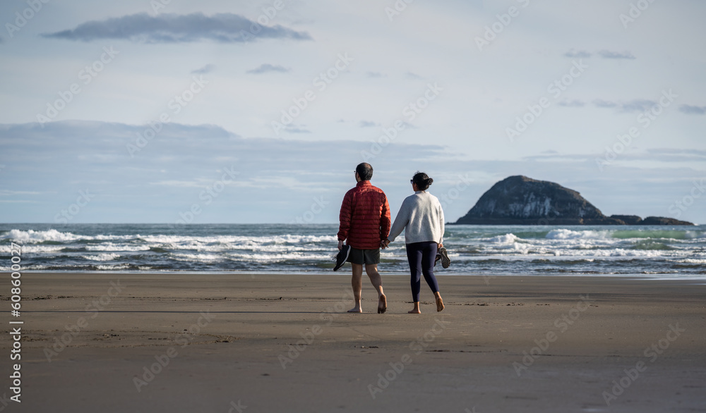 Young couple holding hands and walking barefoot on Muriwai beach. Oaia Island in the distance, Auckland.