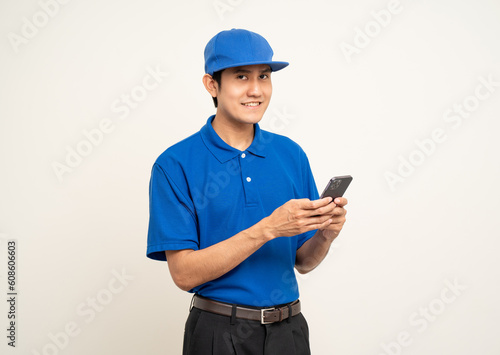 Asian man in blue uniform standing holding smartphone on isolated white background. Male service worker with cell phone. Delivery courier shipping service