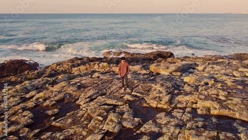 Young Man Standing On The Rocky Cliffs With Rough Sea Waves In Arteixo, La Coruna, Galicia, Spain. Pullback Shot  photo