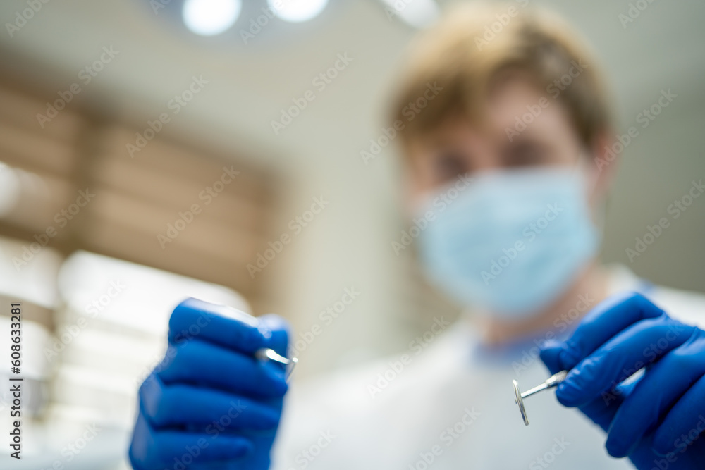Blurred face of blond male dentist in medical uniform wearing blue medical mask and gloves while holding stomatological instruments in dental cabinet Blurred face of blond male dentist in medical uniform wearing blue medical mask and gloves while holding stomatological instruments in dental cabinet