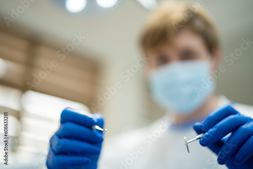 Blurred face of blond male dentist in medical uniform wearing blue medical mask and gloves while holding stomatological instruments in dental cabinet 