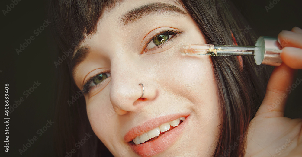 An attractive brunette with a nose piercing applies serum to her face