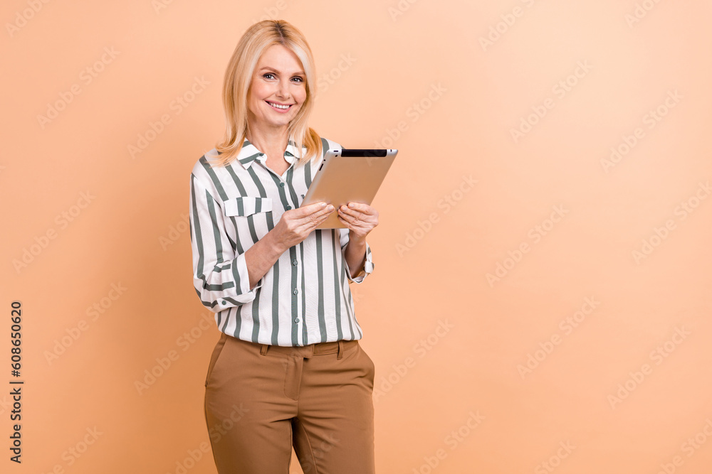 Portrait of positive optimistic retired woman wear striped formalwear hold tablet reads statistics isolated on beige color background Portrait of positive optimistic retired woman wear striped formalwear hold tablet reads statistics isolated on beige color background