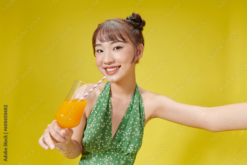 Delightful Asian woman drinking orange juice with a straw while standing against yellow Delightful Asian woman drinking orange juice with a straw while standing against yellow