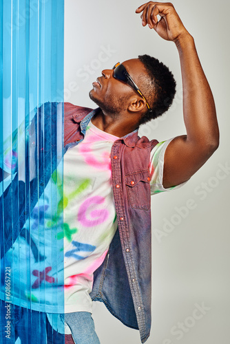 Side view of trendy young afroamerican man in sunglasses, colorful denim vest and t-shirt posing and standing behind blue polycarbonate sheet on grey background, DIY clothing, sustainable lifestyle