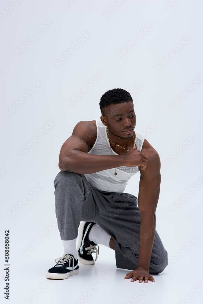 Full length of muscular young african american man in tank top and pants touching golden necklace and posing confidently in stylish and trendy outfit on grey background, good looking Full length of muscular young african american man in tank top and pants touching golden necklace and posing confidently in stylish and trendy outfit on grey background, good looking