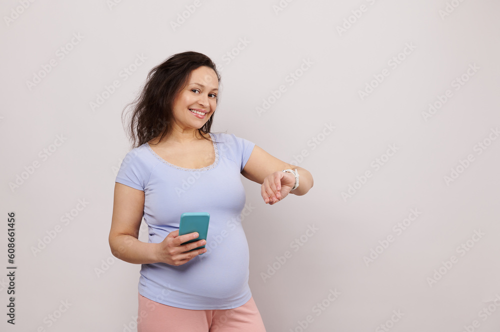 Happy middle aged pregnant woman in 28 week of pregnancy, expecting baby, holding smartphone and checking mobile app on electronic smart wrist watch, smiling looking at camera, over white background Happy middle aged pregnant woman in 28 week of pregnancy, expecting baby, holding smartphone and checking mobile app on electronic smart wrist watch, smiling looking at camera, over white background