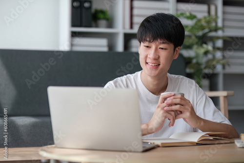 A happy and smart young Asian male college student in casual clothes is doing his homework at a coffee table in his living room.