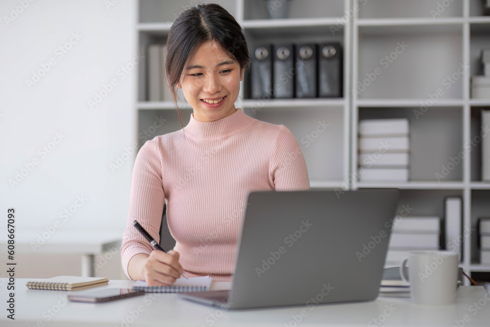 A beautiful young Asian businesswoman focuses on her work, taking notes on notepad and using her laptop computer at her desk in the office. A beautiful young Asian businesswoman focuses on her work, taking notes on notepad and using her laptop computer at her desk in the office.