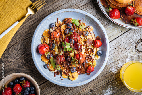 Small American pancakes mixed with seasonal fruit like strawberries, blueberries and cherries. Dipped with melted chocolate, placed on a rustic wooden board. 