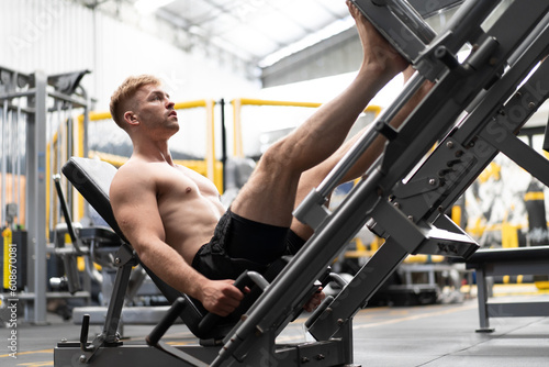 Young muscular man naked torso exercise using press machine to strength his legs in sport gym. Fit man training body workout doing heavy weights leg pushing equipment with quad muscles in fitness club