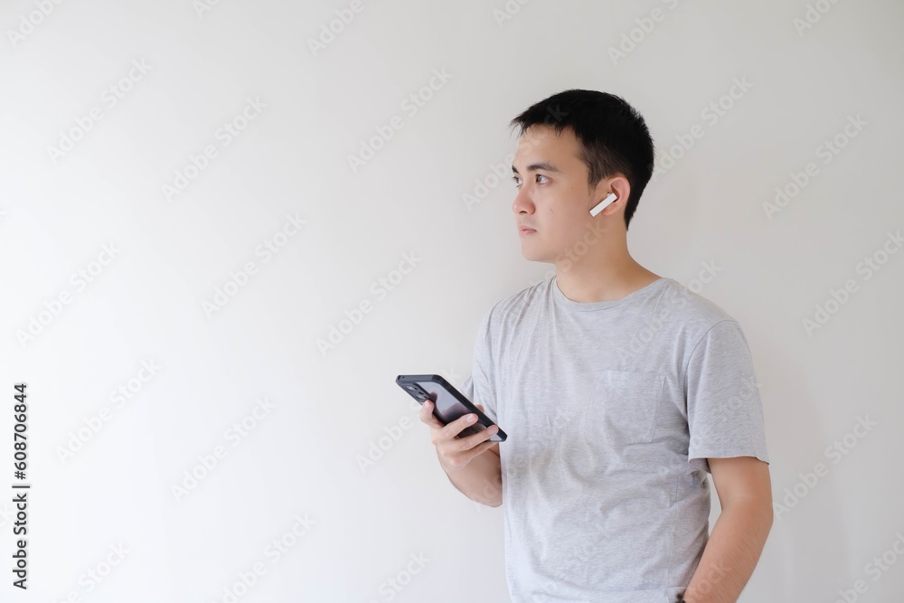 A young Asian man who wears a grey t-shirt, a pair of wireless earphones, and a smartwatch is holding a smartphone and looking at the left side. Isolated white background. A young Asian man who wears a grey t-shirt, a pair of wireless earphones, and a smartwatch is holding a smartphone and looking at the left side. Isolated white background.