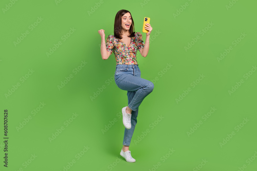 Full length photo of delighted funny person use smart phone raise fist success isolated on green color background Full length photo of delighted funny person use smart phone raise fist success isolated on green color background