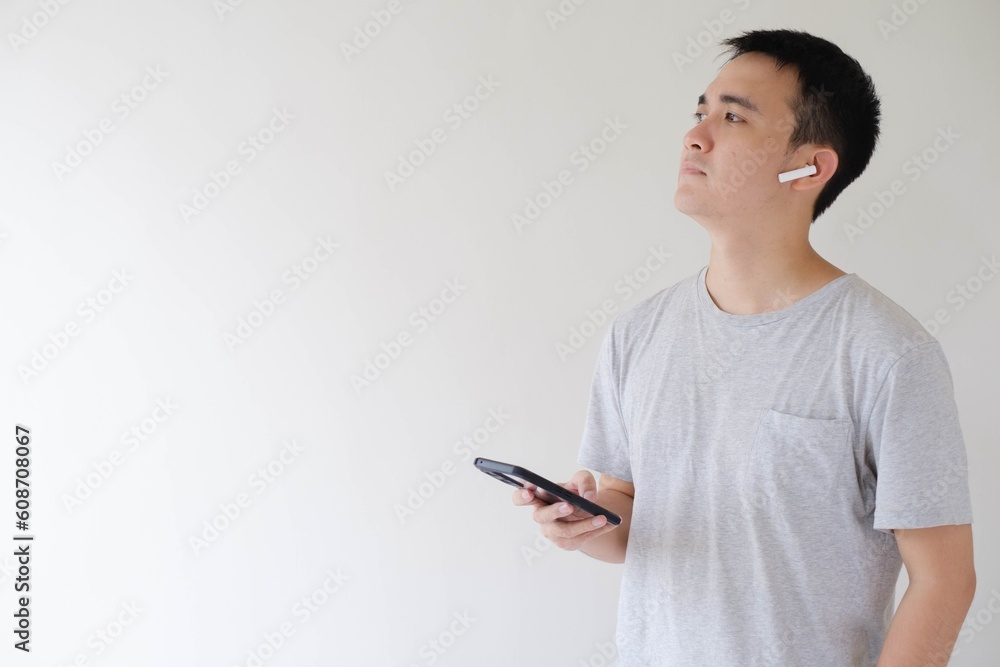 A young Asian man who wears a grey t-shirt, a pair of wireless earphones, and a smartwatch is holding a smartphone and looking at the left side. Isolated white background. A young Asian man who wears a grey t-shirt, a pair of wireless earphones, and a smartwatch is holding a smartphone and looking at the left side. Isolated white background.
