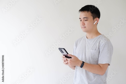 A young Asian man who wears a grey t-shirt, using a pair of wireless earphones and a smartwatch on his left wrist is holding and looking at a smartphone. Isolated white background. © David