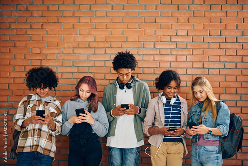 Multiracial group of teenagers using their cell phones while standing against wall.