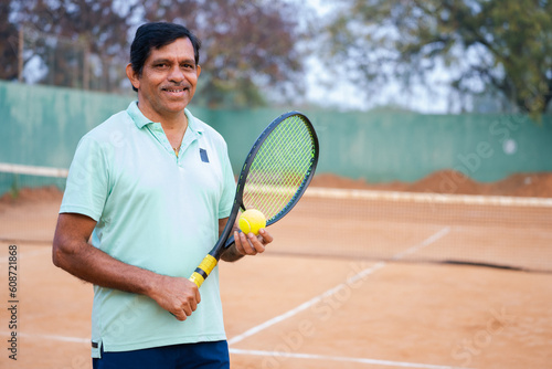 Happy indian senior man with racquet and ball by looking at camera at tennis court - concept of hobbies, successful and relaxation. © WESTOCK