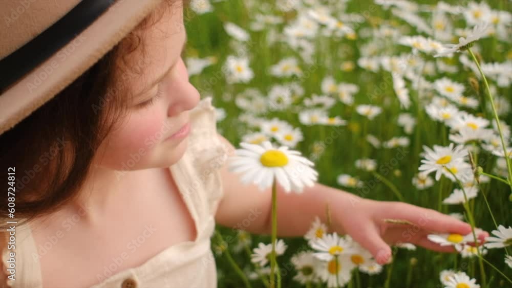 Side view of charming little girl in hat sitting on meadow enjoys smell chamomiles at summer day, laughing and smiling. Happy pretty child plays with flowers outdoors. Nature and childhood concept Side view of charming little girl in hat sitting on meadow enjoys smell chamomiles at summer day, laughing and smiling. Happy pretty child plays with flowers outdoors. Nature and childhood concept