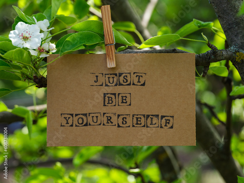 Piece of cardboard with words Just Be Yourself on it hanging on a pear tree branch with blossoms and leaves using a wooden clothespin. © rosinka79