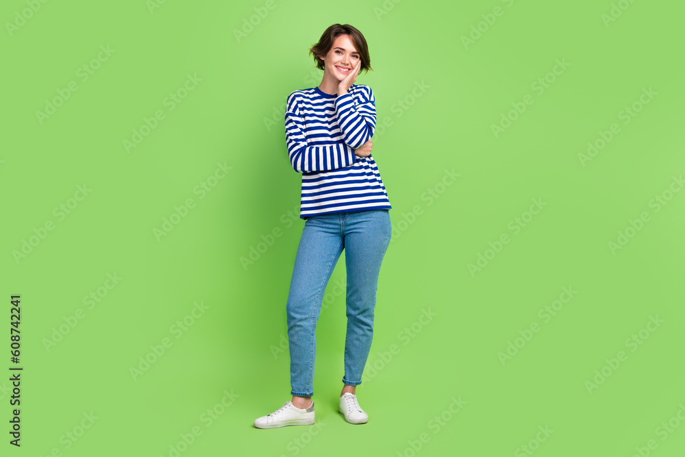 Full length photo of good mood adorable woman wear sailor shirt denim trousers hold palm on cheekbone isolated on green color background Full length photo of good mood adorable woman wear sailor shirt denim trousers hold palm on cheekbone isolated on green color background