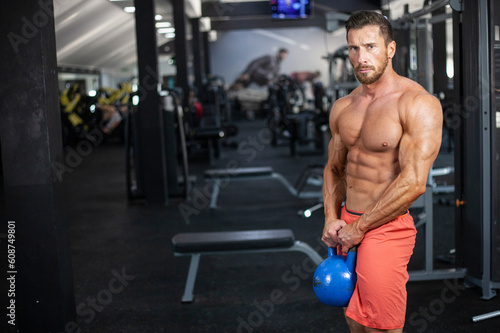 Strong , powerful, muscular man holding blue kettlebell with both hands in a gym