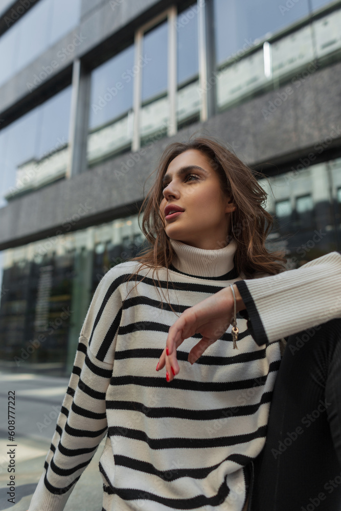 Pretty fashion glamour lady in stylish striped sweater sits on the street near a modern building