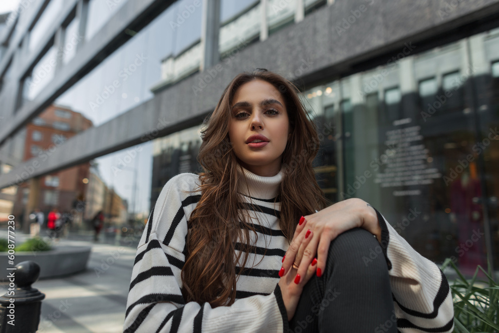 Adorable fashion pretty woman in stylish striped sweater sits in the city near a modern building