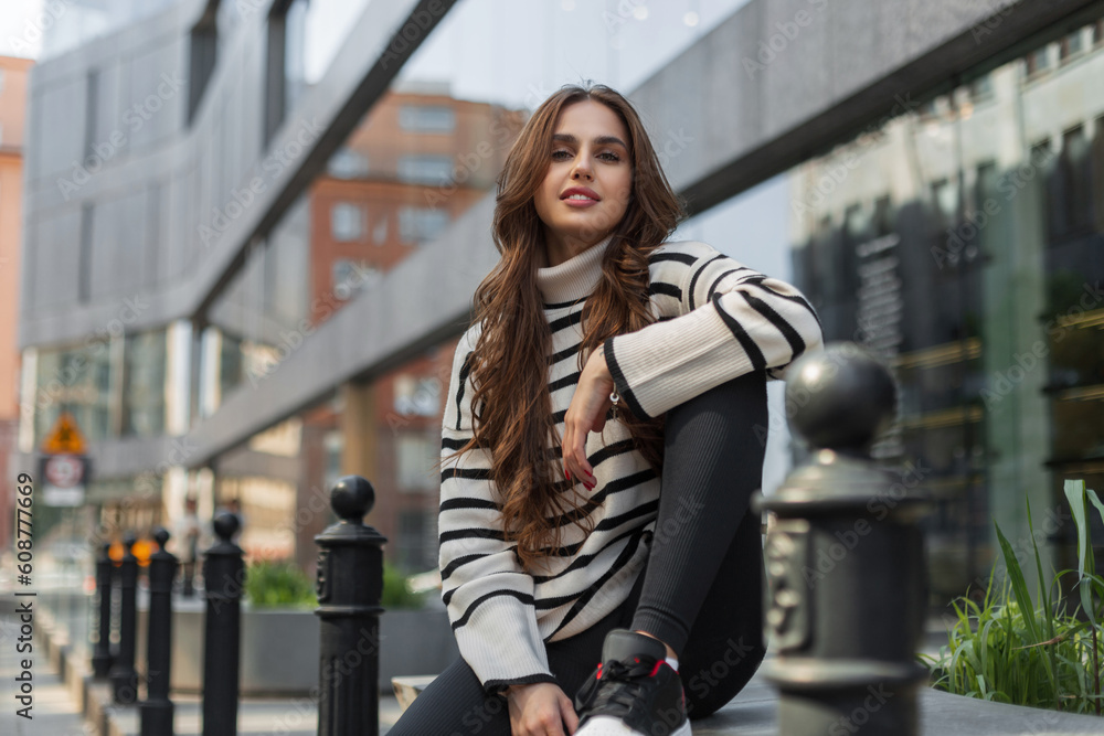 Happy beautiful young fashionable woman model in casual stylish clothes with a striped sweater and black leggings sits in the city near the modern mall