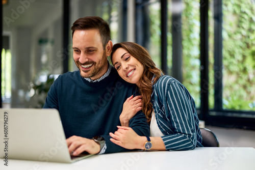 A smiling female employee hugs her male partner while he works on a laptop.
