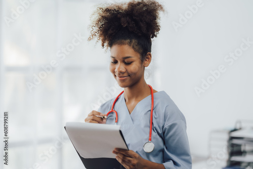 Young African american female doctor using smartphone and working with computer while sitting at the clinic room.