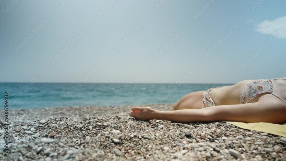 A young woman in a bikini is lying on a yellow towel by the sea on a rocky beach, with a blue sky. A young woman in a bikini is lying on a yellow towel by the sea on a rocky beach, with a blue sky.