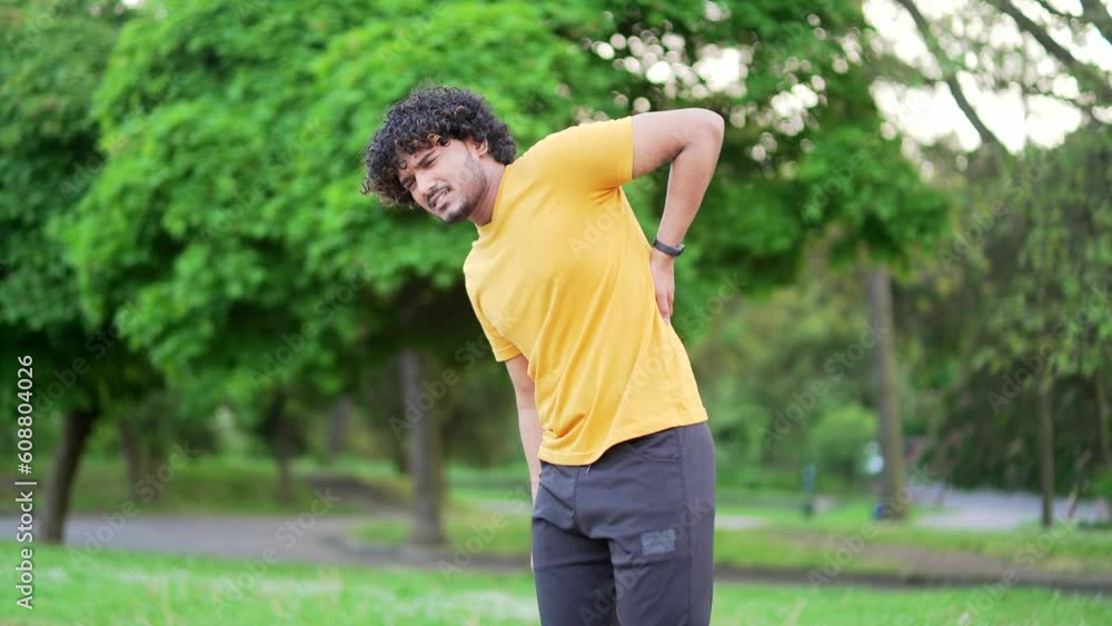 Young adult runner suffering from back pain, sports injury, muscle spasm in urban city park. Handsome man in a T-shirt having back flank ache and problem after exercise outside, massaging a sore spot