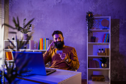 A happy entrepreneur is sitting in a neon blue and purple lighted home office with a credit card in his hands and talking on the phone.