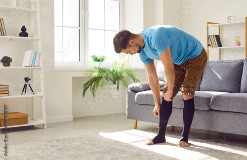 Bild auf Leinwand Young man standing by the sofa in the living room and putting elastic stretchabl