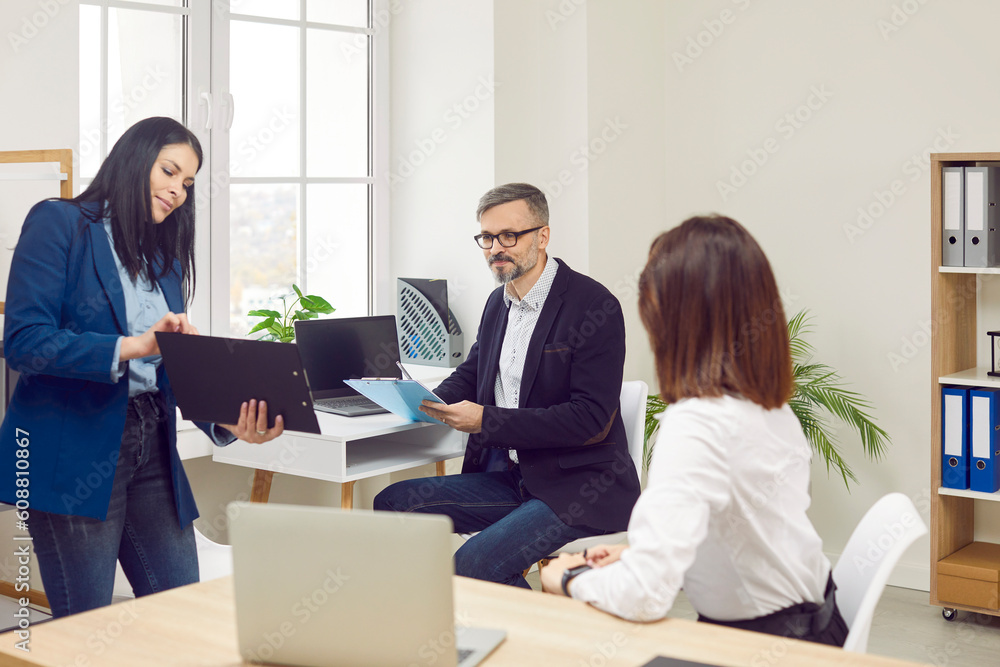 Young woman employee showing something to her colleagues on the workplace in the office. Three coworkers discussing a new projects or startups, analyzing company. Team work concept. Young woman employee showing something to her colleagues on the workplace in the office. Three coworkers discussing a new projects or startups, analyzing company. Team work concept.