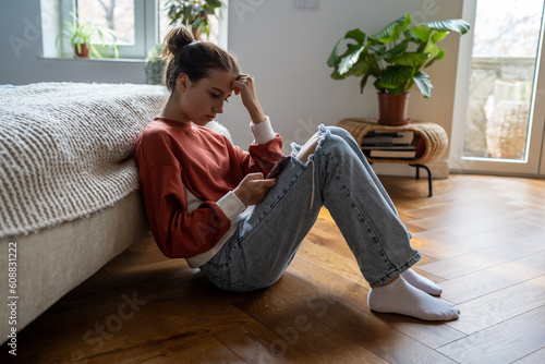 Teens and cyberbullying. Upset teen girl sitting on floor near bed using smartphone at home, scrolling social media. Child spending too much time on phone. Teenagers and gadget addiction photo