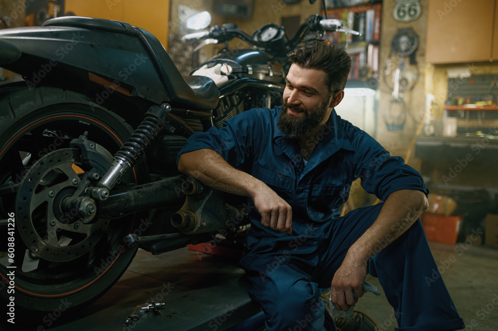 Satisfied smiling mechanic feeling glad looking at repaired motorcycle Satisfied smiling mechanic feeling glad looking at repaired motorcycle