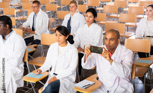 Portrait of interested hispanic doctor attending refresher course, using smartphone to record lecture