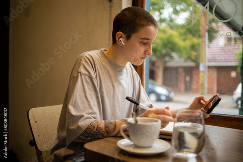 Latin transgender generation z student using mobile phone, digital tablet at the bar. He is using earphones for video call 