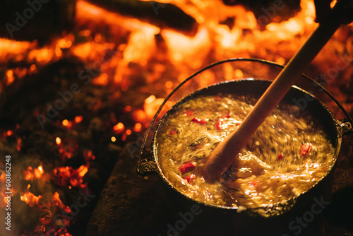 A wooden ladle stirs a pot of peppery soup in a cast iron pot over red hot coals