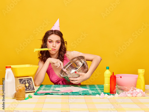 Young girl with bright makeup and birthday cap cooking cake for her party against vivid yellow background. Mess and chaos. Concept of party, celebration, emotions, female beauty, youth. Pop art photo