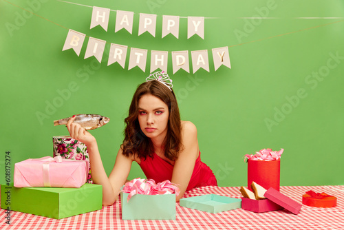 Young birthday girl with calm, serious face holding fish against green background. Weird, crazy presents. Concept of party, celebration, emotions, female beauty, youth. Pop art photo