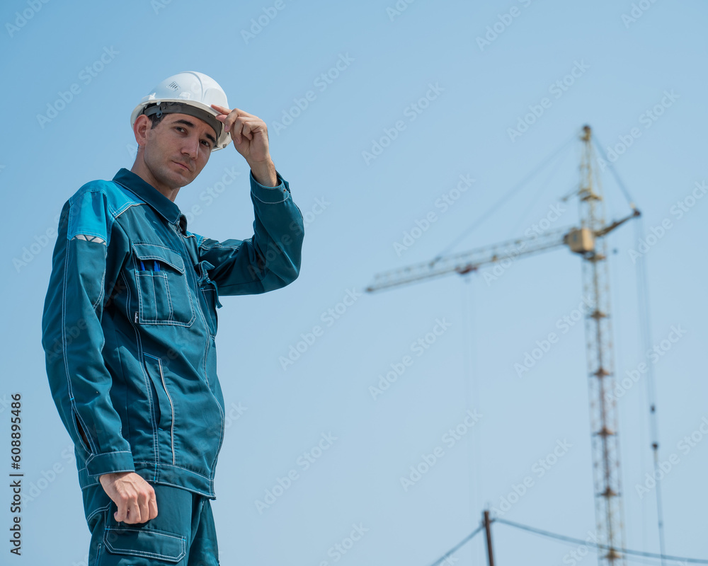 A builder in work clothes and a helmet stands on a construction site against the background of a construction crane. A builder in work clothes and a helmet stands on a construction site against the background of a construction crane.