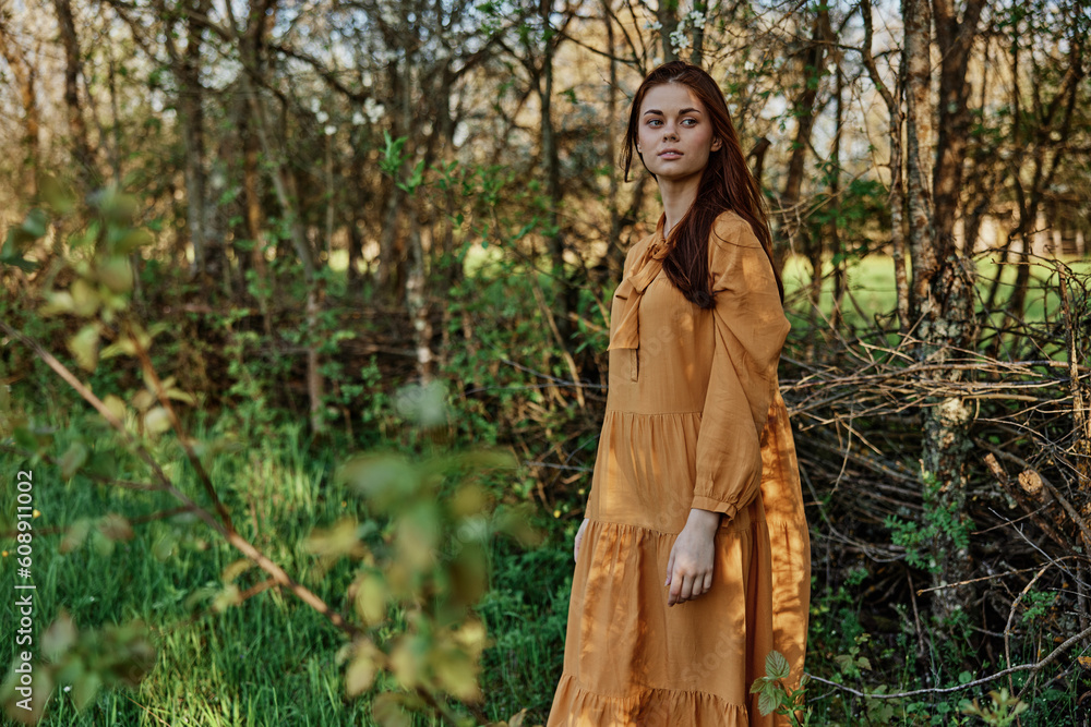 a woman with long hair walks in the shade near the trees, dressed in a long orange dress, enjoying the weather and the weekend, while looking at the camera. The theme of privacy with nature a woman with long hair walks in the shade near the trees, dressed in a long orange dress, enjoying the weather and the weekend, while looking at the camera. The theme of privacy with nature