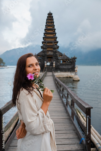 - [x] Portrait of a young European girl, brunette, with brown eyes, travels to the sights of Bali, posing against the backdrop of a Balinese temple on the water, holding a flower in her hand.