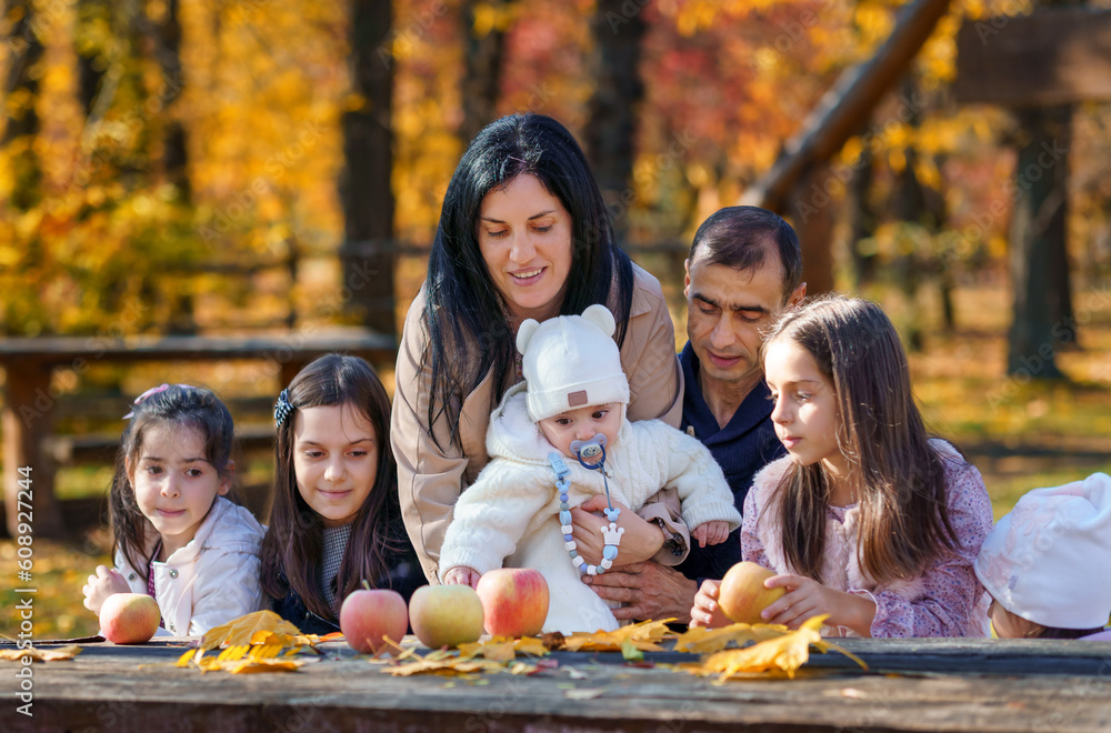 Fototapeta premium big family has picnic in autumn city park, children and parents sitting together at the table, with apples and yellow maple leaves, happy people enjoying beautiful nature, bright sunny day