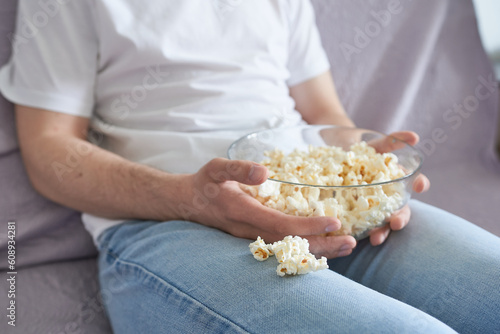 Young man with bowl of popcorn sitting on sofa at home, closeup