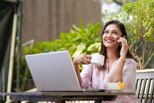 Indian woman drinking tea or coffee, using laptop and talking on smartphone.